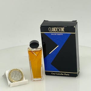 Puede incluir: Una botella en miniatura de "Clandestine" Eau de Toilette de Guy Laroche Paris. El frasco de perfume es rectangular con un tapón negro, lleno de líquido ámbar. La caja es negra con un diseño geométrico azul y letras doradas.