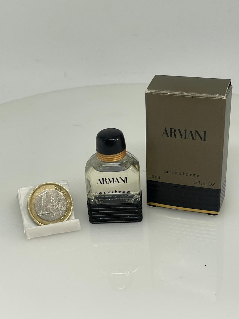 Puede incluir: Un peque&ntilde;o frasco de vidrio transparente de Armani Eau de Toilette para hombre. El frasco tiene una tapa negra y una etiqueta negra con las palabras "Armani" y "eau pour homme" impresas en blanco. El frasco est&aacute; al lado de una peque&ntilde;a caja con el mismo dise&ntilde;o y las palabras "Armani" y "eau pour homme" impresas en blanco. La caja tambi&eacute;n tiene las palabras "10 ml" y "33 FL OZ" impresas en ella.