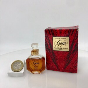 May include: A small, octagonal perfume bottle with a clear, faceted stopper and a gold label that reads "Gem de Van Cleef & Arpels Paris." The bottle is next to a red box with the same branding. A coin is next to the bottle.