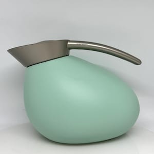 Puede incluir: Una tetera verde menta en forma de huevo con tapa y asa grises. El asa es curva y de metal. La tetera tiene el texto "Georg Jensen" en el asa.