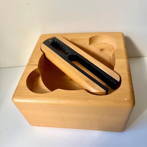 Puede incluir: Un cascanueces de madera con un mecanismo de metal negro descansa dentro de una caja de madera de color claro. El cascanueces tiene un acabado de madera natural con detalles en negro. La caja tiene dos cortes curvos y forma cuadrada.