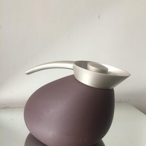 Puede incluir: Una jarra térmica de color morado con tapa y asa plateadas. La jarra tiene una forma redondeada, similar a una piedra.