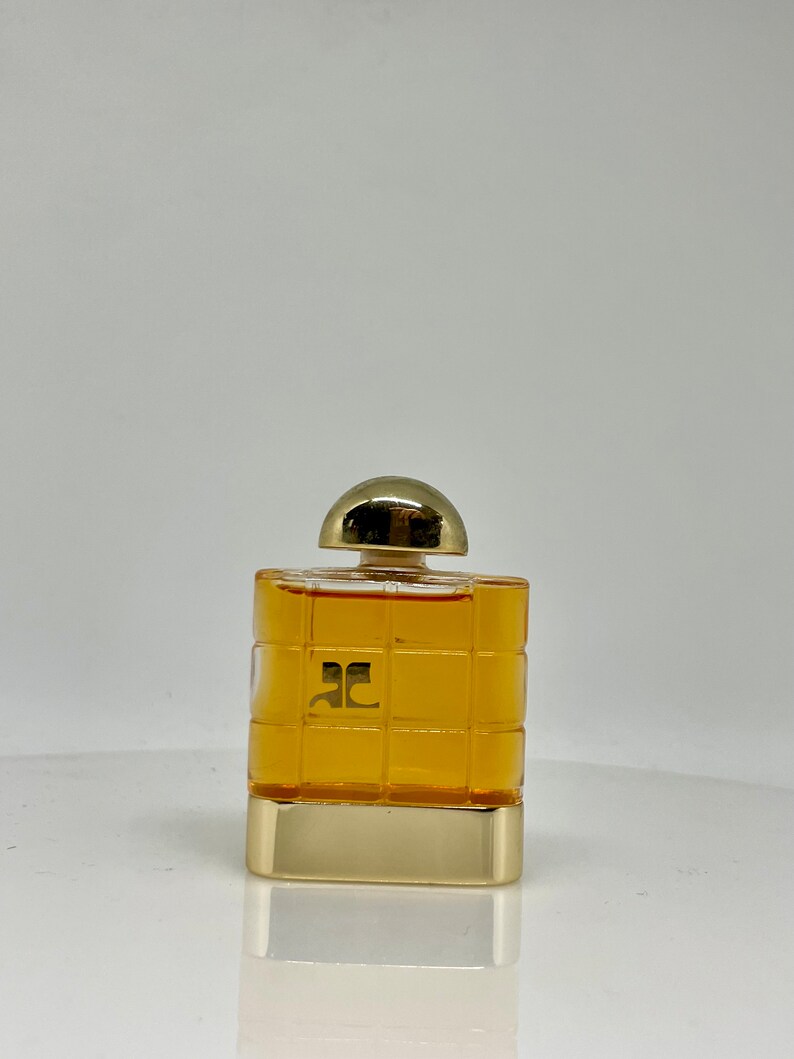 Peut inclure: Un petit flacon de parfum carr&eacute; avec un bouchon et une base dor&eacute;s. La bouteille pr&eacute;sente un motif quadrill&eacute; et contient un liquide de couleur dor&eacute;e. Le logo "ac" est visible sur le devant.