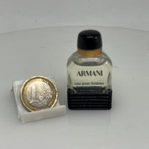 Puede incluir: Un peque&ntilde;o frasco de colonia Armani de vidrio transparente con tapa negra y banda dorada. El frasco est&aacute; sobre una superficie blanca junto a una moneda de euro.