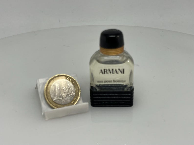 Puede incluir: Un peque&ntilde;o frasco de colonia Armani de vidrio transparente con tapa negra y banda dorada. El frasco est&aacute; sobre una superficie blanca junto a una moneda de euro.