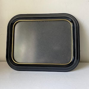 May include: A rectangular black tray with rounded corners and a gold-colored inner trim. The tray has a matte black interior and is set against a white background. The tray is likely used for serving or display.