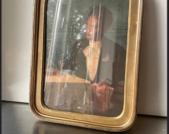 May include: A gold-colored picture frame with a glass front and a portrait of a person in a tuxedo. The portrait is set against a dark green background.