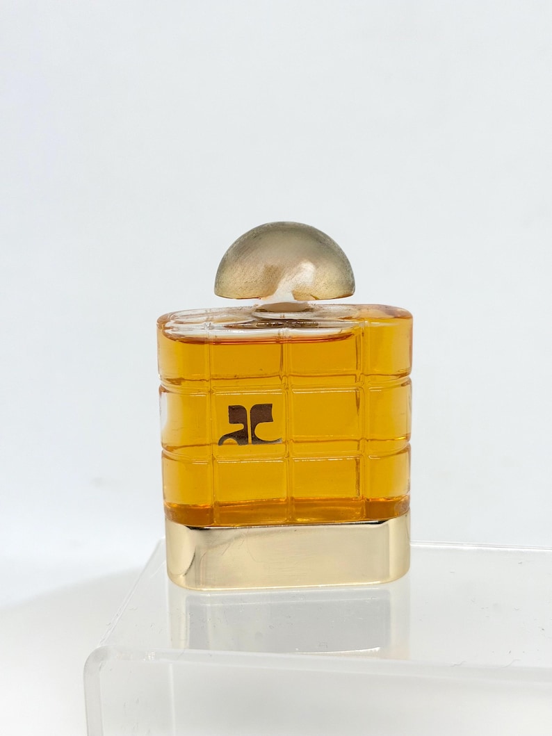 Peut inclure: Un petit flacon de parfum carr&eacute; avec une base dor&eacute;e et un bouchon transparent en forme de d&ocirc;me. Le flacon est de couleur ambre et pr&eacute;sente un motif quadrill&eacute;. Un logo noir est visible &agrave; l'avant.