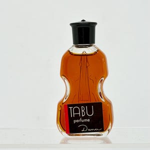 May include: A vintage Tabu perfume bottle with a violin-shaped design. The bottle contains a golden-brown liquid and has a black cap. The label reads "TABU perfume Dana" in white text on a black and red background.
