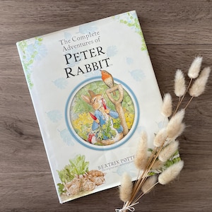 May include: A white book with a blue border titled "The Complete Adventures of Peter Rabbit" by Beatrix Potter. The cover features an illustration of Peter Rabbit holding a carrot and a robin perched on a spade.