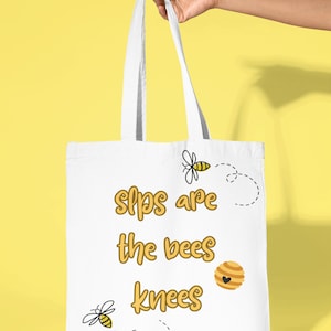 May include: White tote bag with the text "Slips are the bees knees" in yellow lettering. The bag has two bee illustrations and a honeycomb illustration.