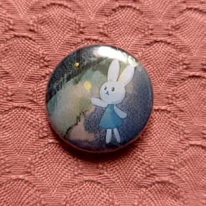 May include: A round button featuring a cartoon rabbit in a blue dress, set against a night sky with stars and a landscape. The button is on a textured pink surface.