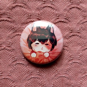 Può includere: Un bottone rotondo con un'illustrazione di un cartone animato di un animale dai capelli marroni su uno sfondo rosa e bianco. L'animale tiene un oggetto rosa e bianco.