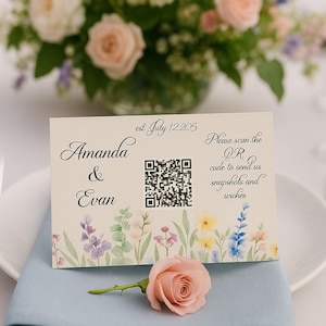 以下が含まれることがあります： 結婚式のテーブルセッティング。花のアレンジメントと「Amanda & Evan」とQRコードが書かれたカード。水色のナプキンとピンクのバラが皿の上にあります。カードには「QRコードをスキャンして、スナップショットと願いを送ってください」と書かれています。