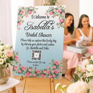 Può includere: Un cartello su un cavalletto dorato recita "Benvenuti all'addio al nubilato di Isabella". Il cartello è decorato con rose rosa e foglie verdi. Il testo sul cartello chiede agli ospiti di condividere foto e auguri. È incluso un codice QR.