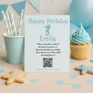May include: A light blue birthday sign with the text "Happy Birthday Emily" and a seahorse illustration. The sign includes a QR code and instructions to upload photos. A cupcake with blue frosting, paper straws, and starfish cookies are also visible.