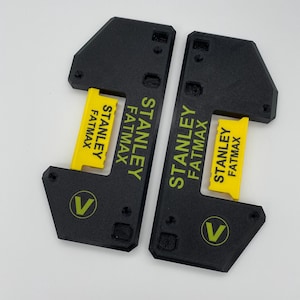 Op de afbeelding: Twee zwarte plastic gereedschappen met gele en groene labels met de tekst "Stanley Fatmax". Elk gereedschap heeft een zwarte "V" in een cirkel aan de zijkant.