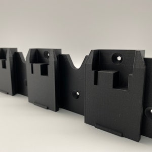 Può includere: Un supporto da parete in plastica nera con tre fessure per tenere oggetti. Le fessure hanno un bordo rialzato in alto e in basso per aiutare a mantenere gli oggetti in posizione.