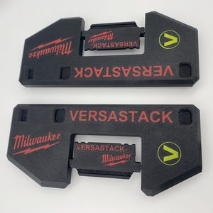 Może przedstawiać: Dwa czarne plastikowe organizery narzędzi VERSASTACK z czerwonymi logami Milwaukee i żółtym logo V. Organizery mają miejsce na narzędzie i miejsce na baterię.