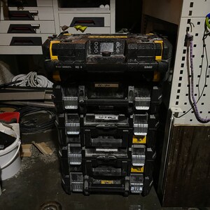 May include: A stack of black toolboxes with yellow accents. The top box has a digital display and the text "DEWALT" on the side. The second box has the text "AV-12M" on the front. The third box has the text "FLD IN 40MM" on the front.