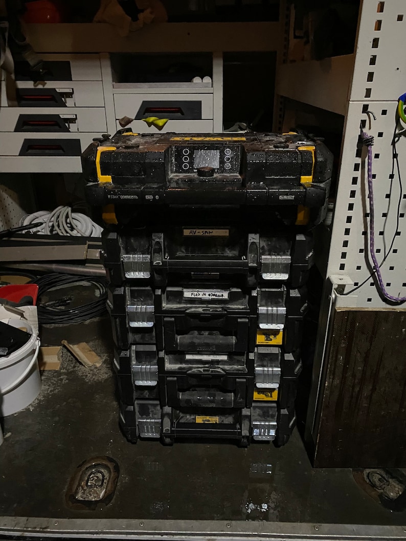 May include: A stack of black toolboxes with yellow accents. The top box has a digital display and the text "DEWALT" on the side. The second box has the text "AV-12M" on the front. The third box has the text "FLD IN 40MM" on the front.