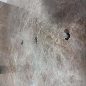 Op de afbeelding: Een close-up van een marmeren muur met drie ronde, zilverkleurige haken. De muur heeft een beige en bruin gemarmerd patroon. De haken zijn gelijkmatig verdeeld en aan de muur bevestigd.