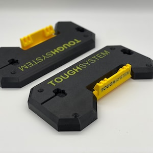 Może przedstawiać: Dwa czarne i żółte uchwyty na narzędzia ToughSystem. Słowo "TOUGHSYSTEM" jest wydrukowane na żółto na każdym uchwycie. Każdy uchwyt ma żółty mechanizm blokujący.
