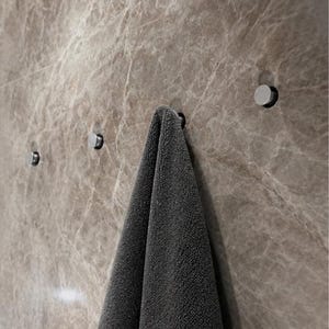 Può includere: Un asciugamano grigio scuro è appeso a uno dei quattro piccoli ganci da parete circolari in metallo argentato. I ganci sono montati su una superficie murale in marmo marrone lucido con venature chiare visibili nella pietra.