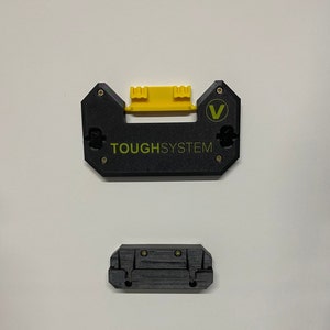 Op de afbeelding: Zwarte kunststof ToughSystem-montagebeugel met een gele kunststof inzet en een groen "V"-logo. De beugel heeft een label met de tekst "TOUGHSYSTEM".