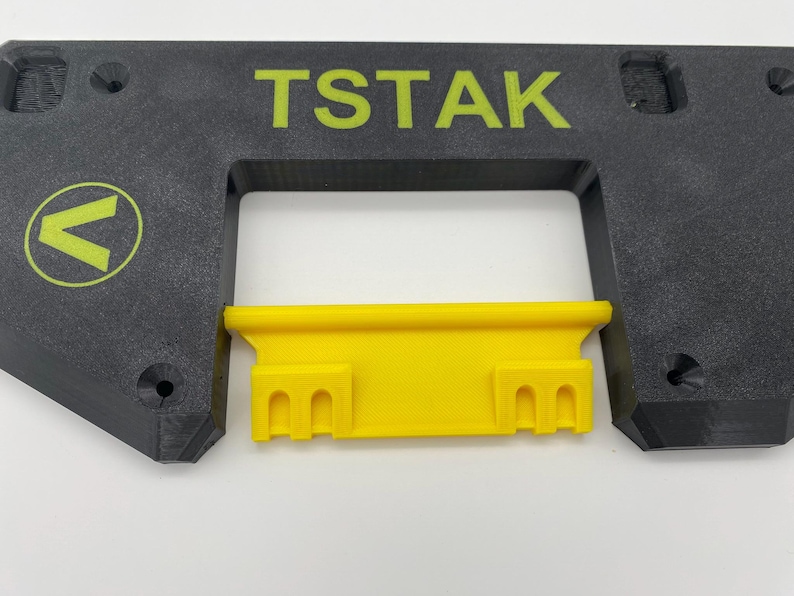 May include: A black plastic 3D printed part with the text "TSTAK" printed in green. A yellow plastic part with a series of holes is inserted into the black part.