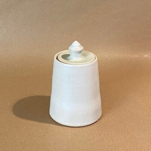 Può includere: Un barattolo di ceramica bianca con un coperchio. Il coperchio ha un piccolo pomo in cima. Il barattolo è su una superficie marrone.