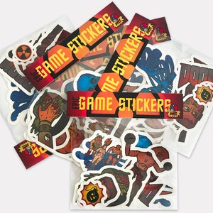 May include: A collection of colorful game stickers in various designs, including characters and symbols. The stickers are packaged in clear plastic and feature the text "GAME STICKERS" in orange and yellow. The stickers are of various sizes.
