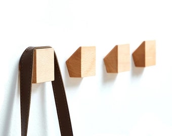 May include: A set of four wooden wall hooks. One hook holds a brown leather strap. The hooks are light-colored wood with a geometric design. The hooks are mounted on a white wall.