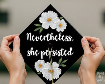 May include: A black graduation cap decorated with white flowers and green leaves. The words "Nevertheless, she persisted" are written in white script. The cap is held by two hands.