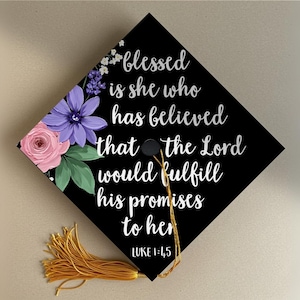 Puede incluir: Gorra de graduación negra decorada con un diseño floral en rosa, morado y blanco. La gorra presenta el texto "blessed is she who has believed that the Lord would fulfill his promises to her" y el versículo bíblico Lucas 1:45.