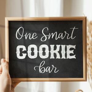 May include: A black chalkboard with a wooden frame, held by a hand. The chalkboard has white chalk writing that says "One Smart Cookie Bar".