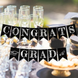 Puede incluir: Pancarta de graduación en blanco y negro con las palabras "Congrats Grad" escritas en fuente de estilo tiza. La pancarta está colgada entre copas de champán y tarros llenos de artículos decorativos.