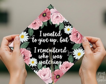 May include: A black graduation cap decorated with pink and white flowers. The cap has the text "I wanted to give up, but I remembered who was watching" written on it.