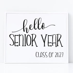 Può includere: Un cartello rettangolare bianco con testo nero che recita hello SENIOR YEAR e CLASS OF 2027. La parola hello è scritta in un carattere corsivo, mentre SENIOR YEAR e CLASS OF 2027 sono visualizzati in un carattere sans-serif semplice e pulito.