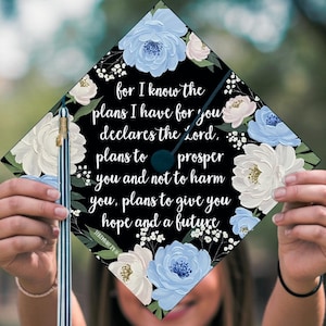 Puede incluir: Un gorro de graduación negro con un diseño floral con flores azules y blancas. El gorro tiene una cita de Jeremías 29:11: "Porque yo sé los planes que tengo para vosotros, declara Jehová, planes de bien y no de mal, para daros un futuro y una esperanza."
