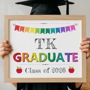 Puede incluir: Un letrero enmarcado de graduación con el texto "TK GRADUATE Class of 2026". El letrero tiene una pancarta de arcoíris y gráficos de manzanas. El marco es de madera clara.