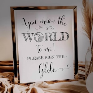 May include: A gold-framed sign with the text "You mean the WORLD to me! - Please sign the Globe" in black cursive font. The word "WORLD" is stylized with a hand-drawn globe inside the letter "O".