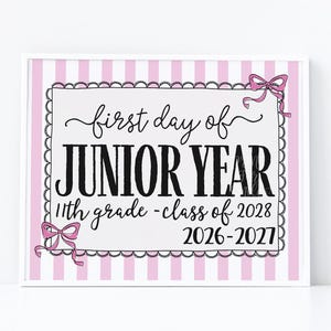 May include: A rectangular sign with a pink and white vertical striped background. The center features a white rectangle with a scalloped black border and two pink bows. The text reads: first day of JUNIOR YEAR 11th grade - class of 2028 2026-2027.