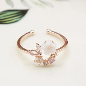 Könnte beinhalten: Ein zarter Goldring mit einer weißen Blume und Perlenakzenten. Der Ring ist verstellbar und hat ein florales Design.