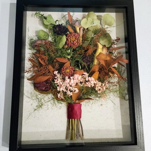 May include: A framed pressed flower bouquet with a burgundy ribbon. The bouquet includes dried flowers in shades of pink, orange, and brown.