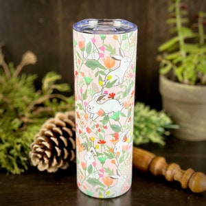 May include: A stainless steel tumbler with a pink background and a pattern of white bunnies and colorful flowers. The tumbler is approximately 20 ounces in size.