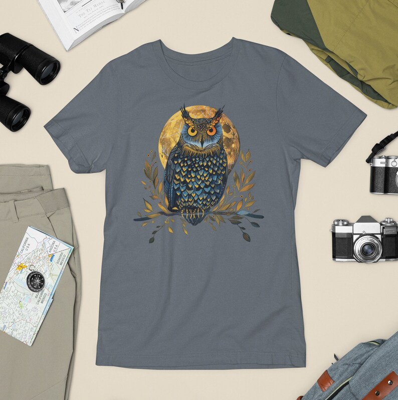 A T-shirt with a blue and yellow illustration of an owl sitting on a branch in front of a full moon. The shirt is steel blue in color.