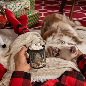 May include: A black ceramic mug with a winter forest scene featuring a Christmas tree, animals, and a red cardinal. The mug is filled with hot chocolate and marshmallows. A dog is lying on a blanket next to the mug.