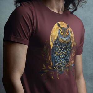 May include: A maroon t-shirt with a graphic of an owl perched on a branch in front of a yellow moon. The owl has blue and black feathers and orange eyes.