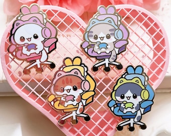 May include: Four enamel pins featuring cartoon cats wearing headphones and sitting in gaming chairs. The cats are pink, gray, orange, and green. The chairs are pink, purple, yellow, and blue. The pins are on a pink heart-shaped grid.